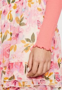 Blommig klänning med mönster av ljusrosa och gula rosor; har en volangkant nedtill. Rosa ribbad långärmad tröja med vågig mudd vid handleden.