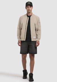 Beige bomberjakke med lynlås foran, elastiske håndled, og sidelommer, båret over en sort t-shirt og parret med mørke shorts og sorte højtop sko.