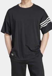 Man med en lös svart t-shirt med tre vita ränder på höger ärm och svarta byxor, med händerna i fickorna.