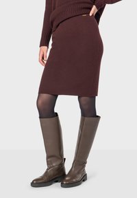 Gonna di maglia bordeaux con silhouette dritta, abbinata a stivali di pelle marroni al ginocchio e collant neri. Presenta un dettaglio in oro.