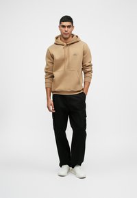 Καφέ φούτερ με μπροστινή τσέπη, κουκούλα με κορδόνια και διακριτικό λογότυπο. Συνδυασμένο με μαύρα cargo παντελόνια και λευκά sneakers.