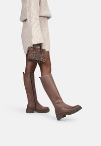 Botas de cuero marrones hasta la rodilla con un acabado texturizado, cremallera lateral y suela robusta. Combinadas con un bolso de piel de leopardo y un suéter ligero.