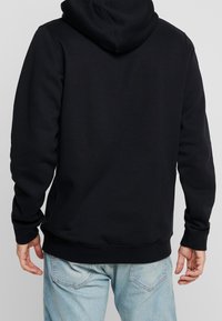 Man som bär en enfärgad svart hooded sweatshirt och ljusblå jeans, sedd bakifrån mot en grå bakgrund.