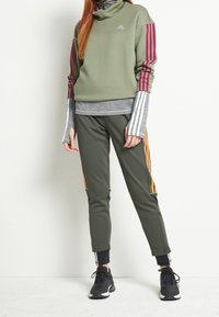 Zielony sweter z bordo paskami, szara warstwa pod spodem i oliwkowe dresy z pomarańczowymi i białymi paskami po bokach. Czarne buty sportowe.