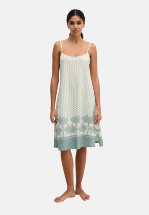 Frau steht barfuß und trägt ein weißes Sommerkleid mit dünnen Trägern, kleinem Muster und einem grünen Palmenmuster am Saum.