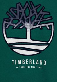 Tejido verde con un logotipo circular blanco que presenta un diseño estilizado de un árbol sobre el texto "TIMBERLAND THE ORIGINAL SINCE 1973" en blanco.