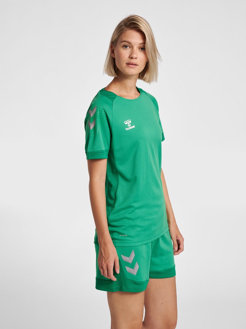 Kobieta stojąca, ubrana w zielony sportowy trykot i pasujące krótkie spodenki z odblaskowymi logo szewronów na rękawie i spodenkach.