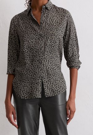 Femme portant une chemise noire à fleurs boutonnée avec les manches retroussées et un pantalon en cuir noir devant un mur blanc.