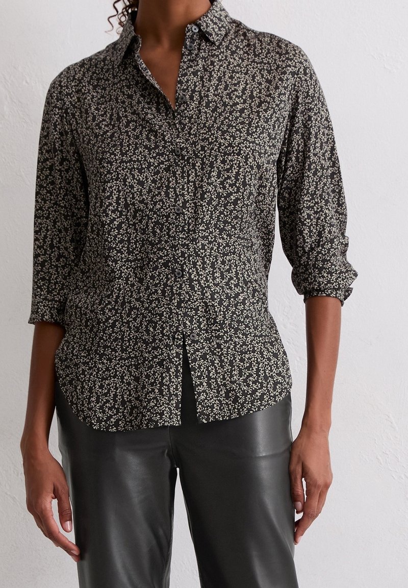 Femme portant une chemise noire à fleurs boutonnée avec les manches retroussées et un pantalon en cuir noir devant un mur blanc.