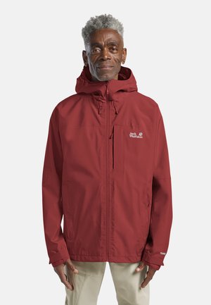 Mann mit kurzen grauen Haaren, der eine rote Jack Wolfskin Kapuzenjacke und beige Hose trägt, steht mit entspannten Händen seitlich am Körper.