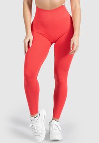 Hoch geschnittene rote Leggings aus dehnbarem Material, mit nahtlosem Design und kombiniert mit weißen Sportschuhen.