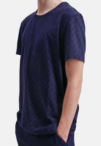 Man in een marineblauwe korte mouwen shirt en bijpassende broek met een subtiele gemêleerde textuur, hand in pocket, tegen een witte achtergrond.