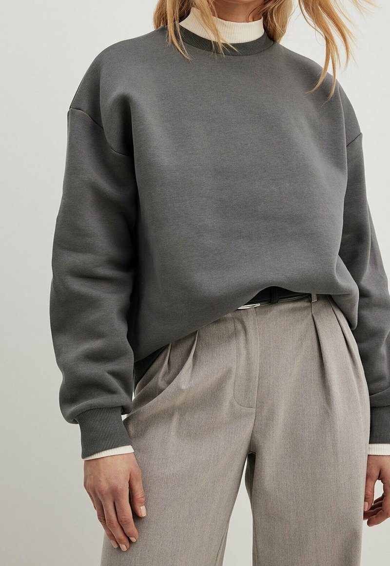Femme portant un sweat-shirt ample gris foncé par-dessus un col roulé blanc, associé à un pantalon beige plissé taille haute et une ceinture noire.