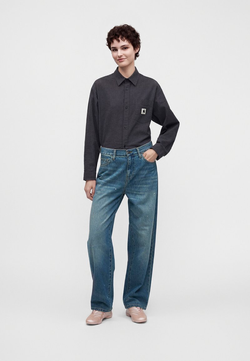 Persona in piedi con una mano in tasca che indossa una camicia grigio scuro con bottoni e jeans larghi blu con scarpe rosa chiaro su sfondo bianco.