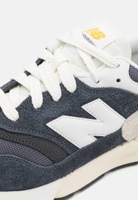 Baskets gris foncé et bleu marine en suède et mesh, avec un logo blanc, des lacets crème et une semelle texturée. Accentuation avec un détail en suède beige.