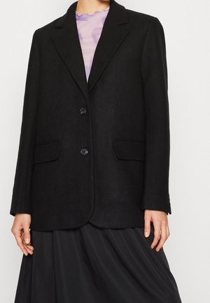 Kobieta ubrana w czarną, dopasowaną marynarkę z dwoma guzikami, kieszeniami z patkami oraz fioletową bluzkę z marszczonym kołnierzem pod spodem.