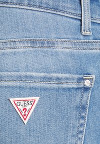 Jeans de mezclilla azul claro con una etiqueta de logo en triángulo roja y blanca "GUESS?" en el bolsillo trasero y detalles de costura sutiles.