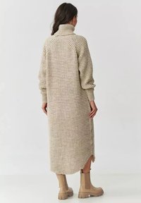 Beżowa dzianinowa sukienka z wysokim kołnierzem, długimi rękawami, teksturowanym wzorem, długości do połowy łydki i bocznymi rozcięciami, zestawiona z beżowymi botkami za kostkę.