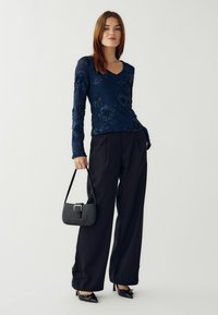 Jūras spēku garās piedurknes tops ar ziedu izšuvumiem, apvienots ar melniem platām kājām biksēm. Rokā tura mazu melnu teksturētu somiņu. Melnas augstpapēžu kurpes.