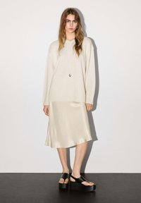 Maglione oversize beige, gonna in crema satinata e sandali con piattaforma neri. Maglia a trama, tessuto liscio e finitura lucida con un design semplificato.
