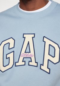 Felpa azzurra chiara con grande logo "GAP" in crema e blu navy, con "Athletic" ricamato in rosa, colletto rotondo, texture in cotone morbido.