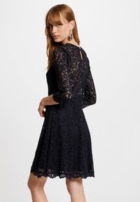 Robe en dentelle bleu marine avec des manches trois-quarts, ourlet festonné, détail en forme de trou de serrure au dos et motif floral en tout. Texture douce.