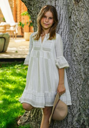 Mädchen in weißem Kleid lehnt an einem Baum, hält einen beigen Hut, draußen auf Gras nahe einer Terrasse mit Korbmöbeln im Hintergrund.