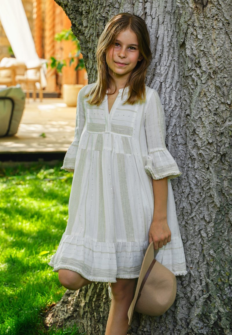 Mädchen in weißem Kleid lehnt an einem Baum, hält einen beigen Hut, draußen auf Gras nahe einer Terrasse mit Korbmöbeln im Hintergrund.