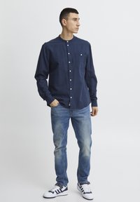 Marineblauwe button-up shirt met een staande kraag en een voorzak, gedragen met lichtblauwe jeans en witte sneakers met marineblauwe accenten.