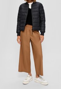 Persona con una giacca a imbottitura nera, sopra nera, pantaloni larghi marroni e sneakers bianche, in piedi contro uno sfondo neutro.
