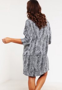 Femme aux longs cheveux bouclés bruns, portant une robe texturée noire et blanche midi, tournée de dos avec un bras légèrement tendu.