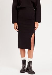 Jupe midi noire en tricot avec une fente latérale, présentant une silhouette ajustée. Associée à des bottes noires et des chaussettes blanches à côtes.