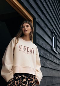 Crèmekleurige sweatshirt met de tekst "SUNDAY" in rood, gecombineerd met een broek met luipaardprint. De stof oogt zacht en de pasvorm is ontspannen met verlaagde schouders.