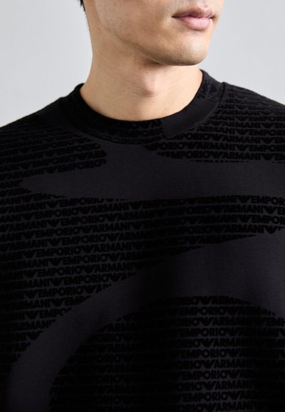 Fekete pulóver, amely az "Emporio Armani" logo texturált mintáival van díszítve. Kerek nyakú, rövidujjú dizájn, puha pamut keverékből készült.