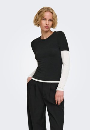 Jeune femme aux cheveux blonds courts portant un haut noir à manches courtes superposé à des manches longues blanches et un pantalon noir ample sur un fond uni.
