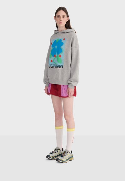 Jeune femme portant un sweat à capuche gris avec un motif de fleurs bleues, un short rouge et rose, des chaussettes blanches montantes avec des rayures jaunes, et des chaussures de course.