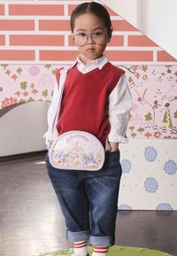 Ungt barn med glasögon, röd väst, vit skjorta, blå jeans och en unicorn-tema axelremsväska som står inomhus med färgglad tapet i bakgrunden.