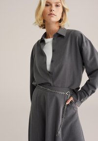 Grå knäppskjorta i ett slätt tyg, kombinerad med en matchande kjol. Har ett silverfärgat kedjebälte med ett hjärtformat detaljer.