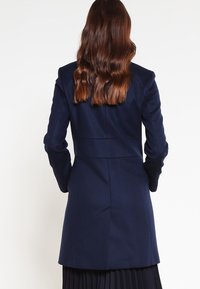 Manteau ajusté bleu marine avec des manches longues, une texture lisse, une silhouette cintrée et une couture de taille définie ; design simple et classique.