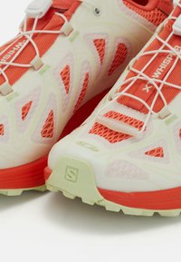 Tenisice za trčanje s gornjim dijelom od mrežice u krem i narančastoj boji, bijelim brzo-podesivim vezicama te rebrastim bež i crvenim potplatima, uz logotipe marki na jeziku i vrhu.