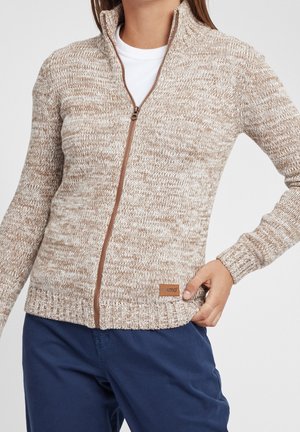 Kobieta ubrana w beżowy i biały melanżowy sweter na zamek, z małą skórzaną metką marki, zestawiony z granatowymi spodniami.