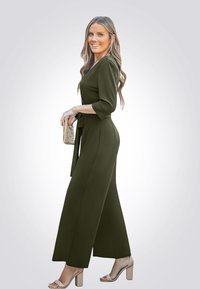 Olivgrüner Jumpsuit mit Taillengürtel, V-Ausschnitt und dreiviertel Ärmeln. Weit ausgestellte Beine, kombiniert mit beigen Riemchen-Heels und einer Clutch.