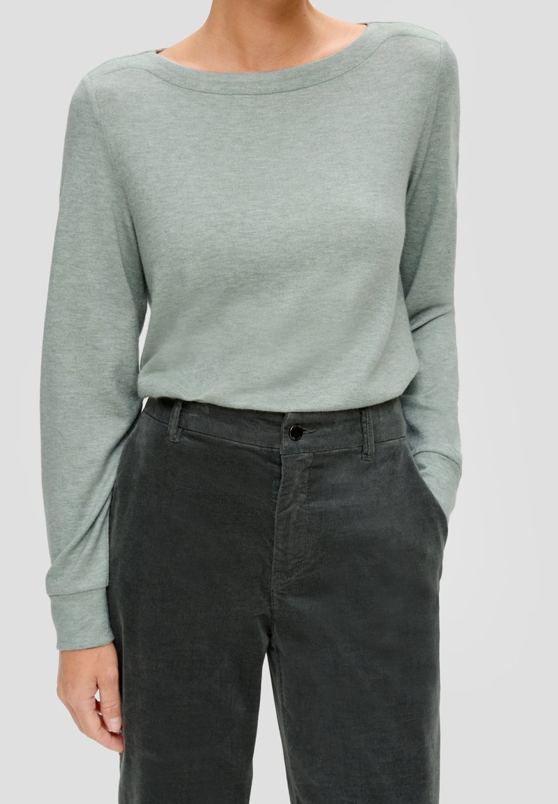 Femme portant un haut à manches longues vert clair rentré dans un pantalon en velours côtelé gris foncé taille haute, les mains dans les poches.