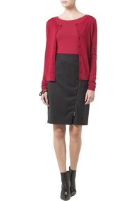 Rode cardigan over een rood shirt, gecombineerd met een zwarte rok met een zijrits. Draagt zwarte enkellaarsjes. Overal gladde texturen.