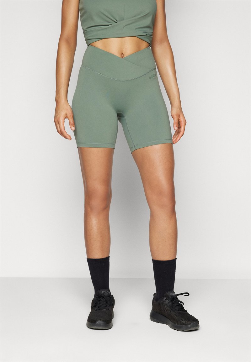 Pantaloni sportivi verde oliva con una cintura alta incrociata. Realizzati in un tessuto elasticizzato, presentano cuciture piatte e una vestibilità aderente. Indossati con sneakers nere.