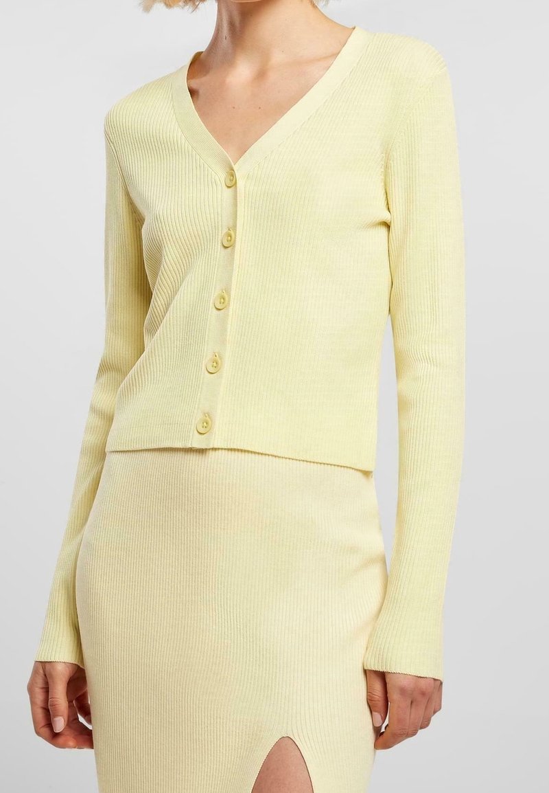 Cardigan a coste di un giallo chiaro con maniche lunghe, scollo a V profondo e sei bottoni sul davanti. Abbinato a una gonna a coste coordinata.