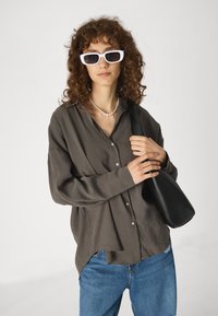 Camisa cinza-escura de botões com um corte descontraído, combinada com jeans azuis e uma bolsa de ombro preta. Óculos de sol retangulares brancos e um colar de pérolas.