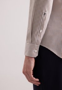 Camicia a quadri marrone e bianca con polsini a bottoni, con dettaglio zip sulla manica sinistra e orlo curvo.
