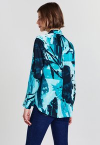 Woman with short brown hair wears abstract blue and black long-sleeve shirt and dark blue jeans, posing sideways on light background.
