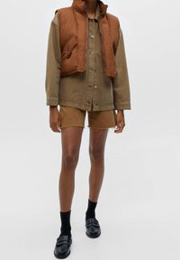 Brown padded vest over a khaki button-up shirt, paired with tan shorts. Black socks and loafers complete the look. Simple, structured design.